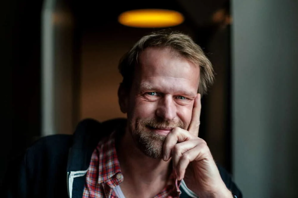 Arne smiling, hand at his face, in warm low light.