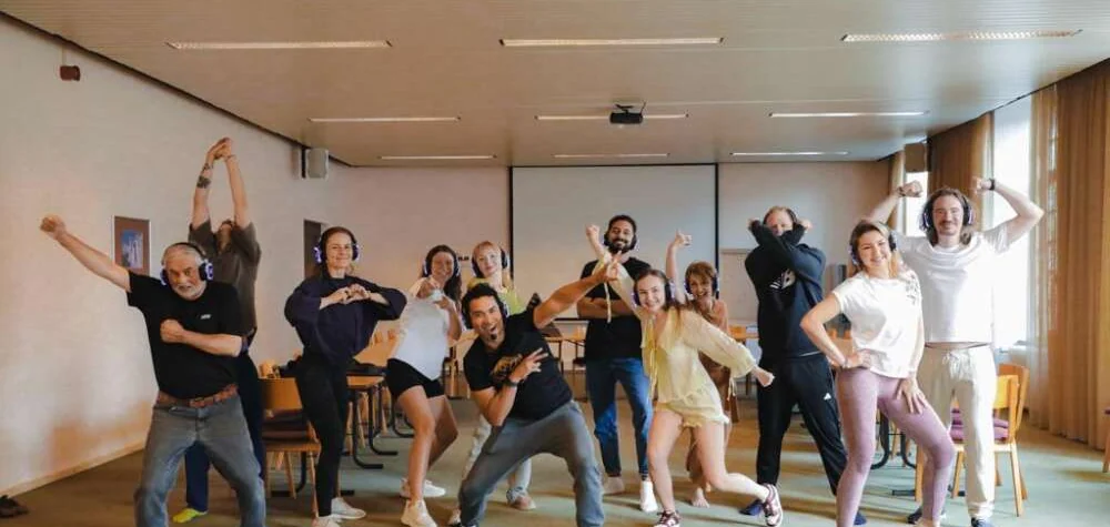 Colleagues in headphones striking playful poses together during a silent-disco team session in a meeting room.