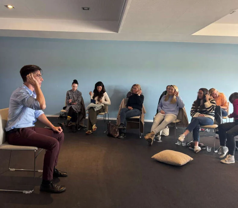A facilitator seated before a circle of colleagues during a workshop in a bright training room.