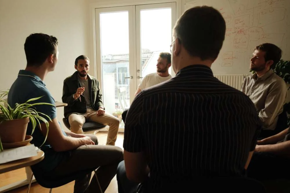 Joshua leading a seated dialogue circle of colleagues in a bright room with a whiteboard.