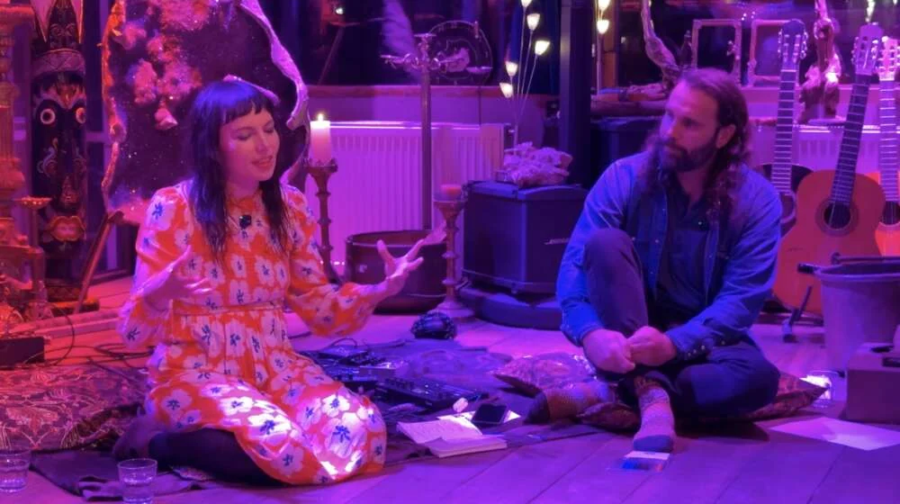 Bloom speaking beside a guest seated on cushions among guitars and candles in a violet-lit room.