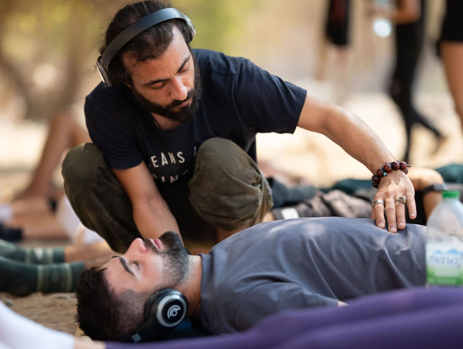 A practitioner kneeling beside a participant lying on a mat, guiding his breathing under trees