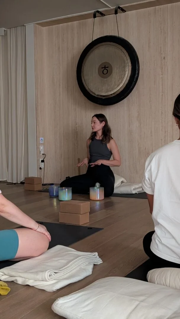 A yoga and sound teacher kneeling before a gong, guiding a group on mats
