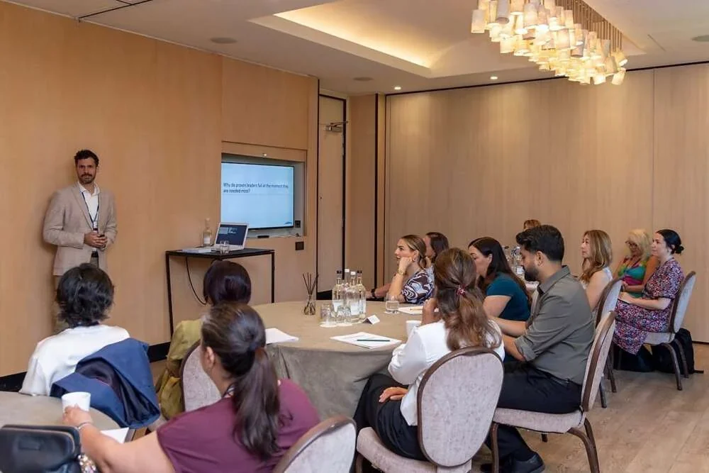 A facilitator presenting to leaders seated around tables in a warm, wood-panelled room
