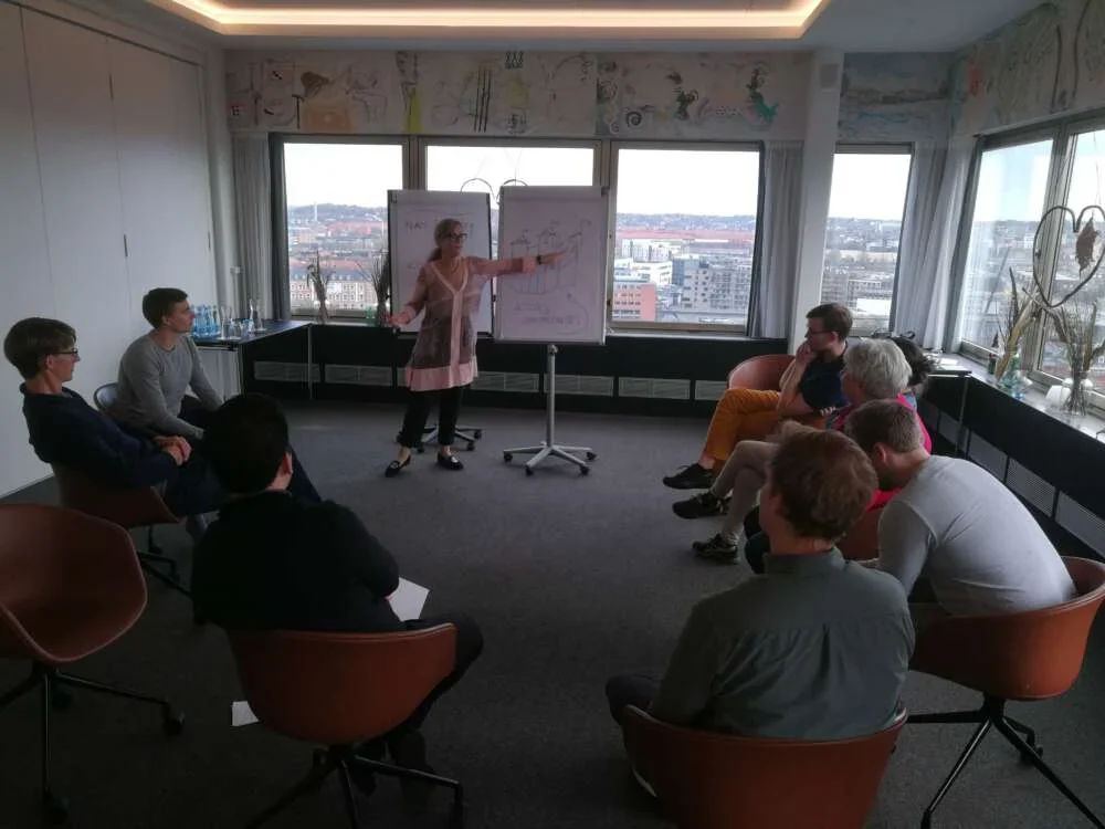 A facilitator drawing on two flip charts for a seated circle of professionals in a high room with city views