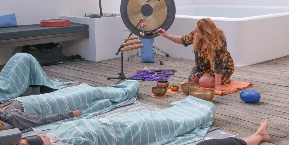 Kitu playing a gong beside singing bowls as colleagues rest under blankets