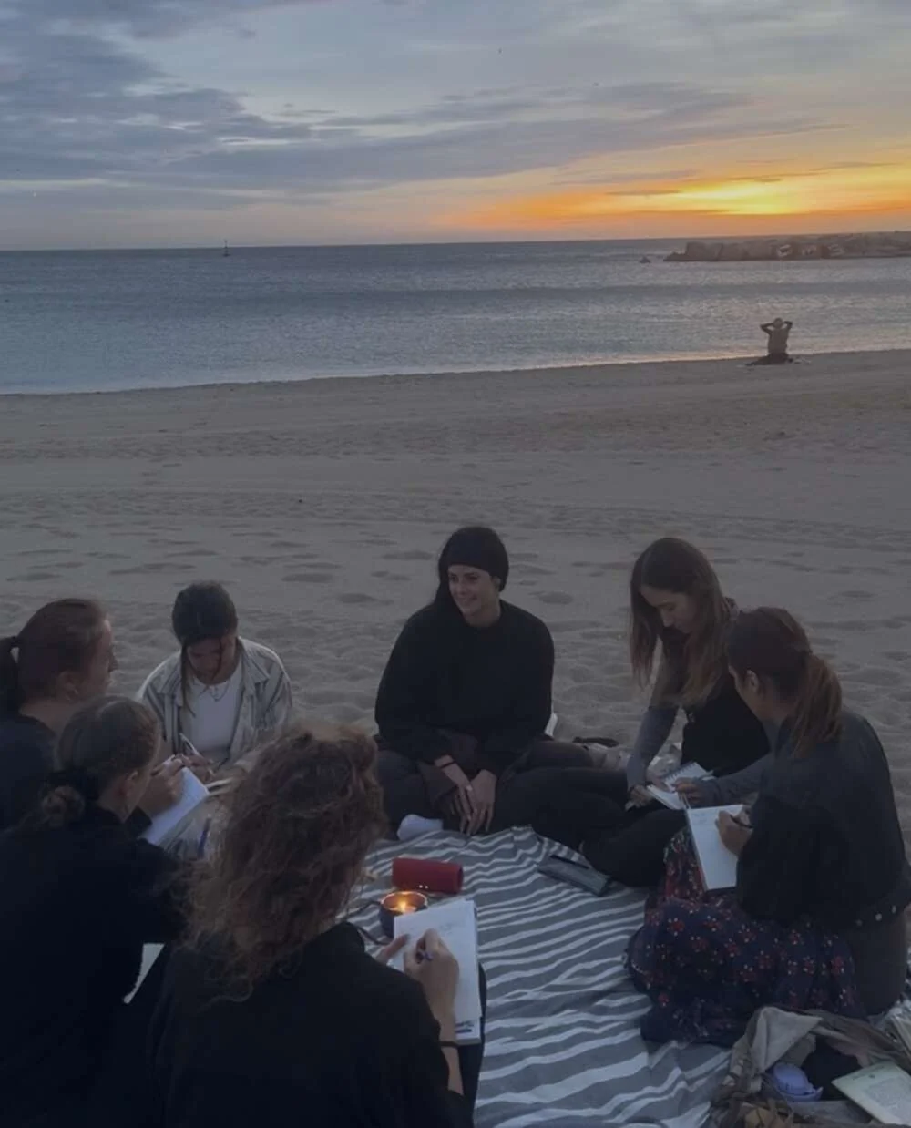 A group writing in journals on a beach at sunset