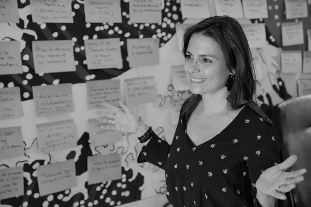 A facilitator beside a wall of notes from a team session, in black and white