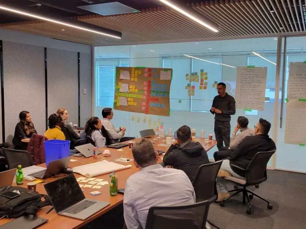 A facilitator standing at the head of a long table as a team works through a session in a glass meeting room lined with sticky notes