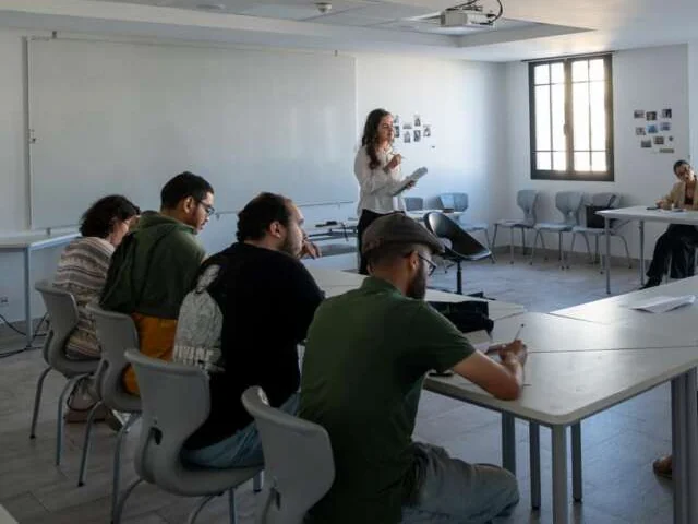 Aicha standing at the front of a bright workshop room as participants write at shared tables