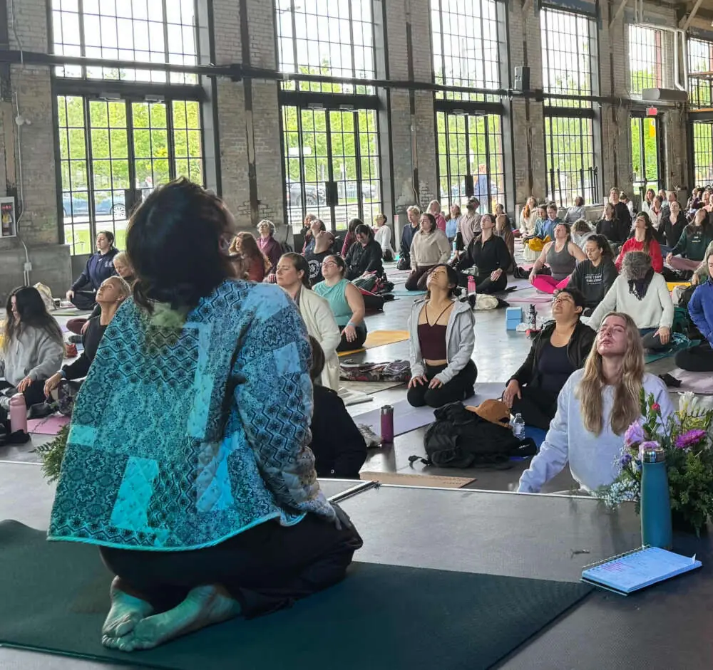 Marianna guiding a hall of people seated on mats through a breathing practice in a converted industrial space