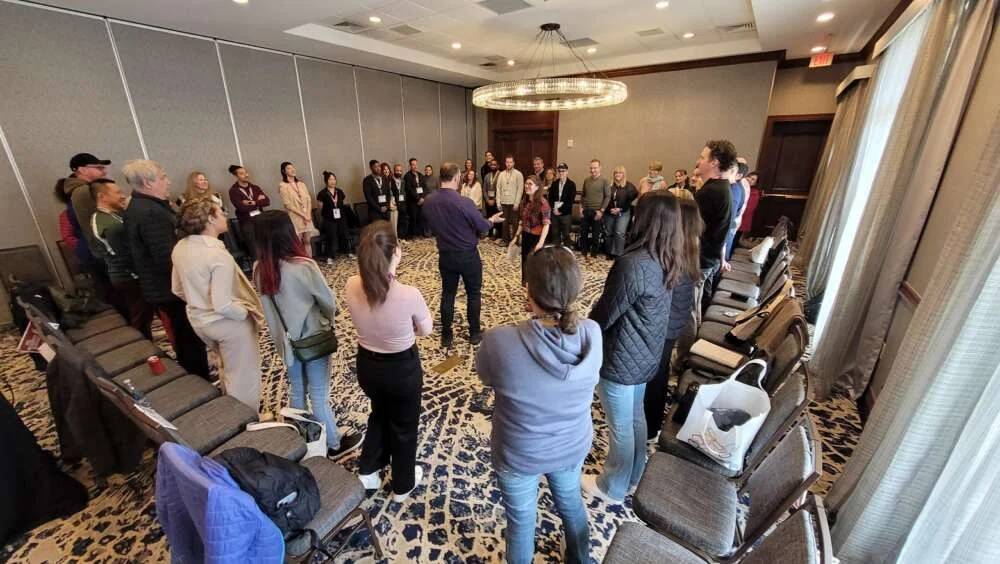 Jordan leading a large standing circle of delegates through an improvisation exercise in a hotel ballroom
