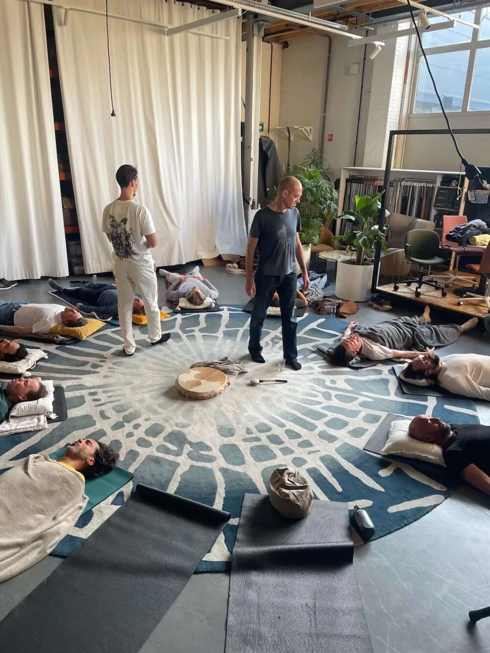A group lying on mats around a frame drum in a bright studio during a guided sound and breath journey