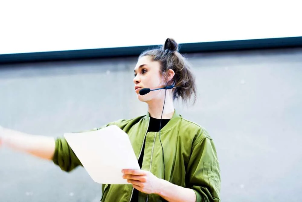 Ida speaking on stage with a headset microphone and her notes