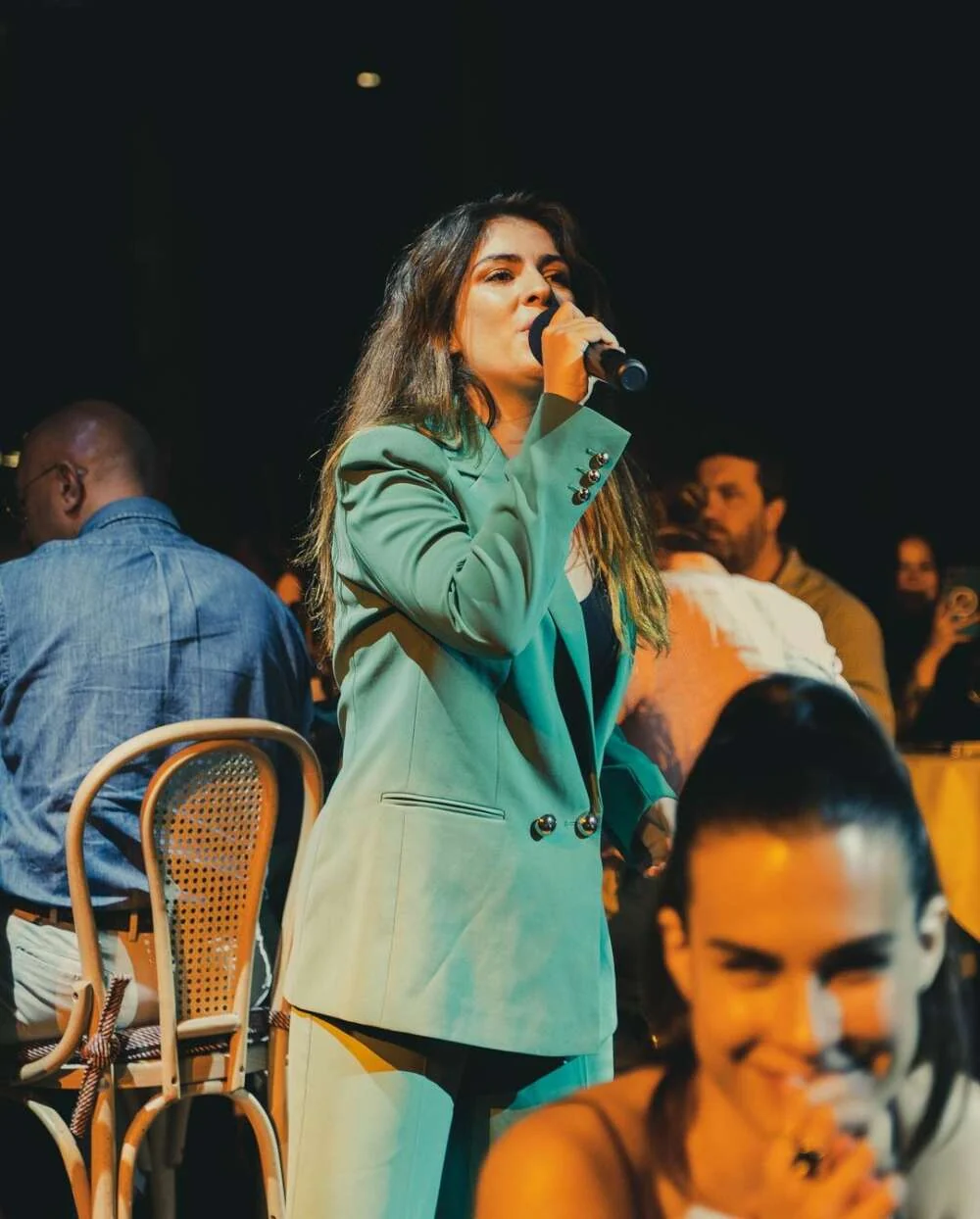 Paola singing into a microphone among guests seated at dinner tables during an evening event