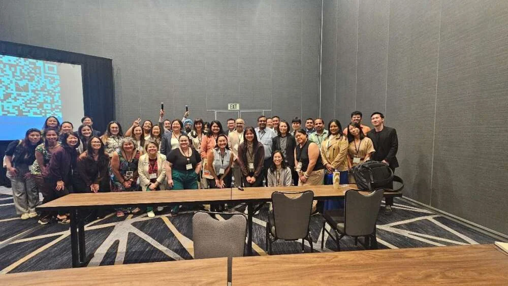 A conference group of about a hundred gathered for a photo in a hotel function room