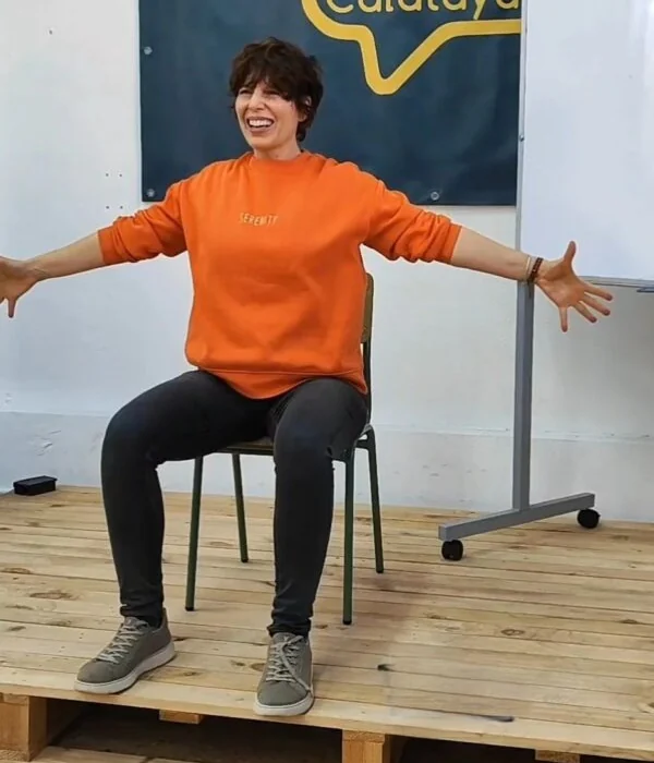 A facilitator laughing with her arms wide, seated on a chair during a movement session at a public school