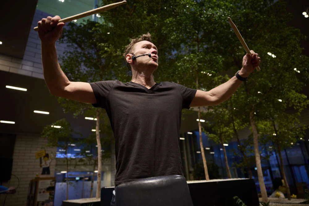 Peter conducting a group with drumsticks raised in a glass atrium.
