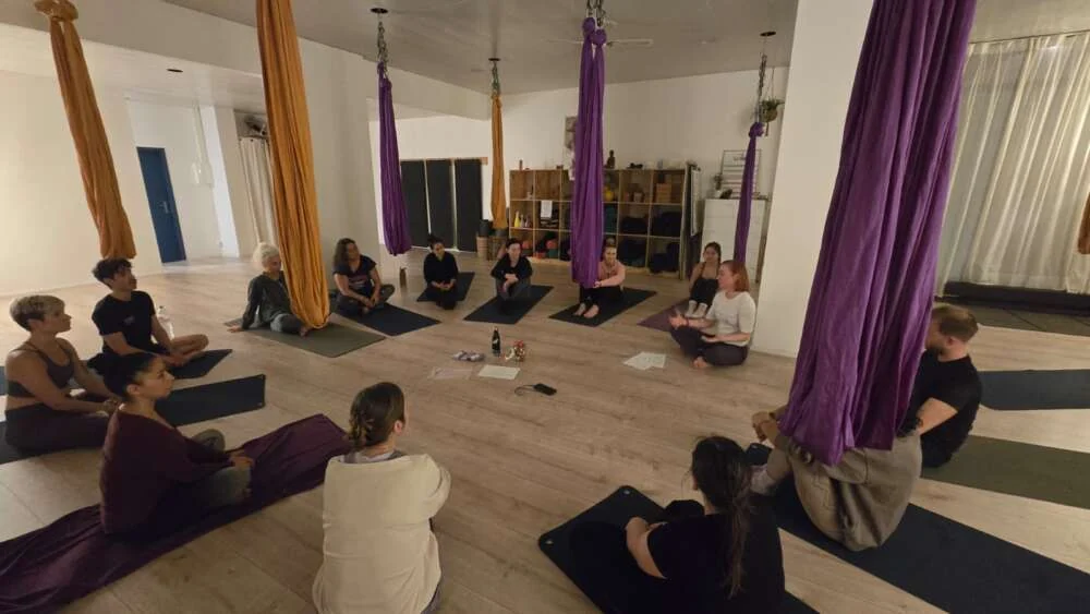 A group resting on mats beneath aerial yoga hammocks in a bright studio