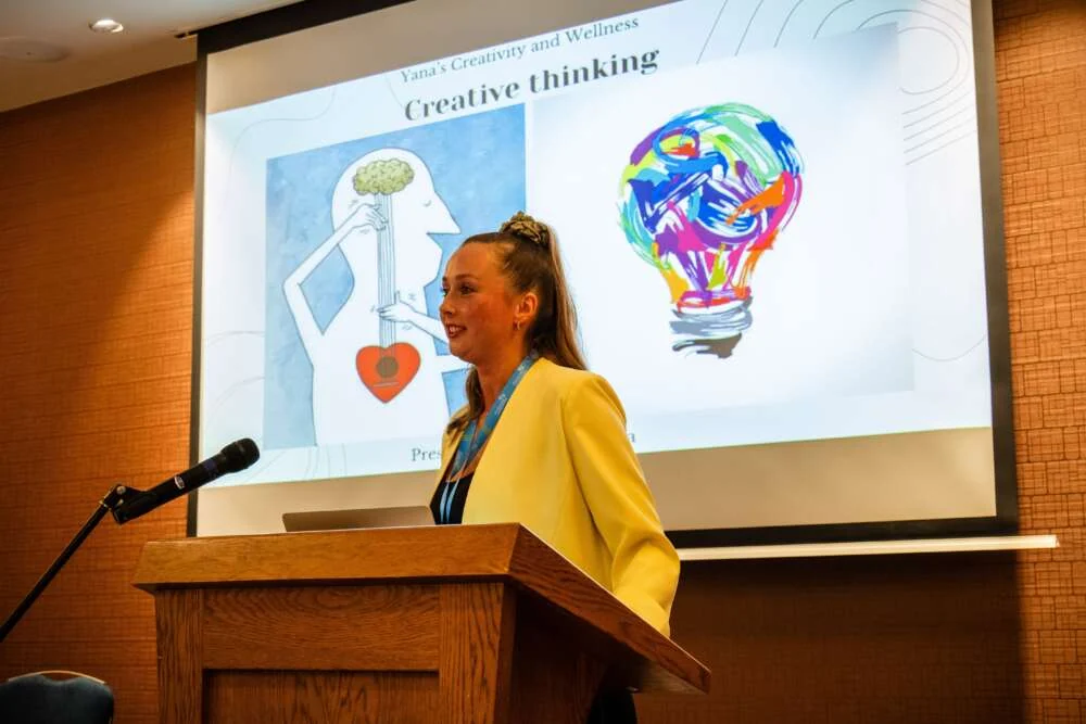 Yana speaking from a lectern in front of a large screen at a conference