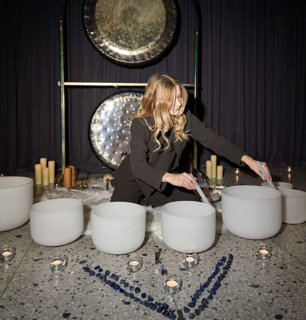 A practitioner plays crystal singing bowls before two gongs, among candles