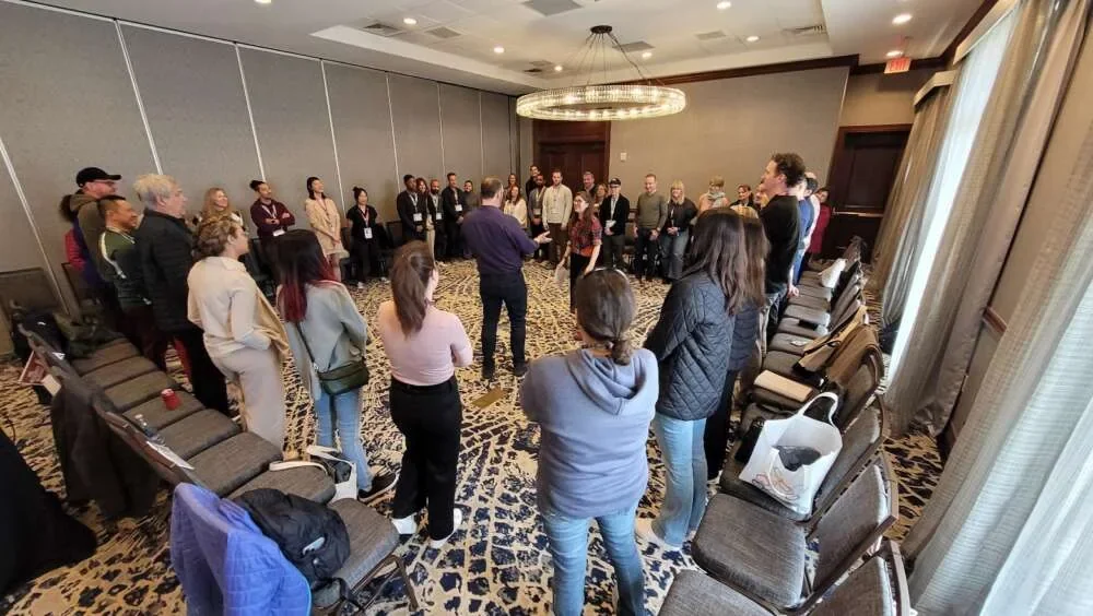 A large group standing in a hotel ballroom during a facilitated improvisation exercise