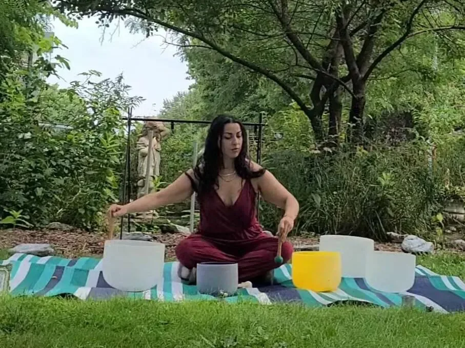 Alexis seated among crystal singing bowls on a blanket in a leafy garden