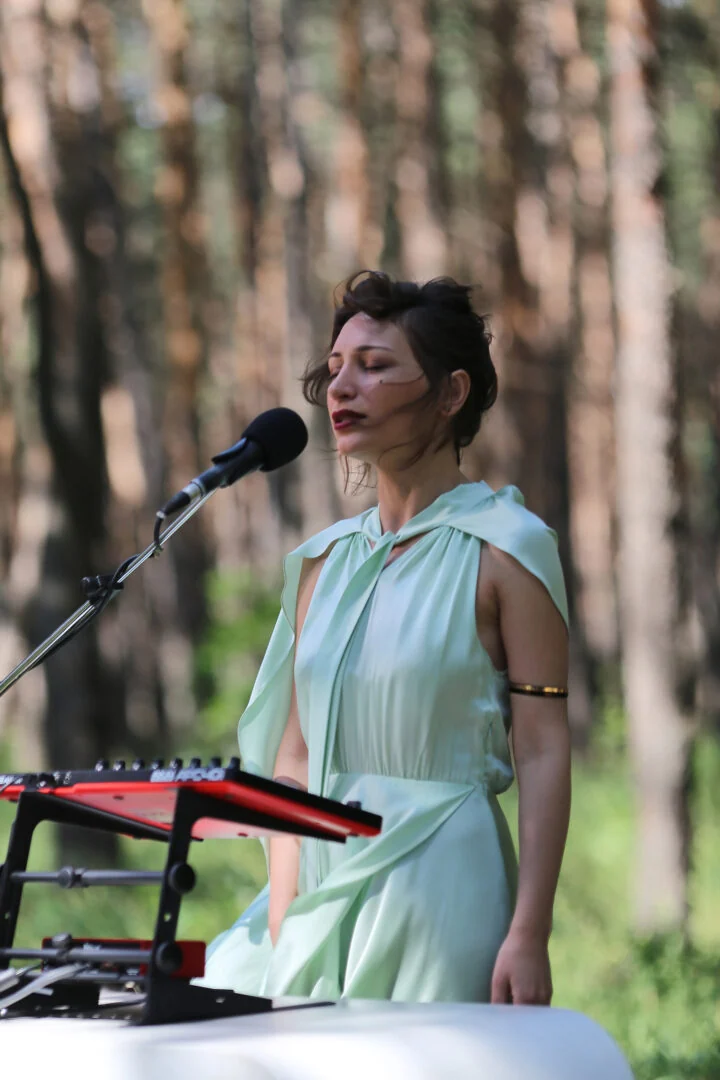 Nuci singing at a microphone beside a keyboard among pine trees