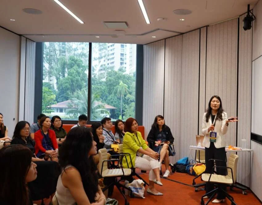 An organisational psychologist leading a workshop for a seated group in a Singapore meeting room, tropical trees outside the window