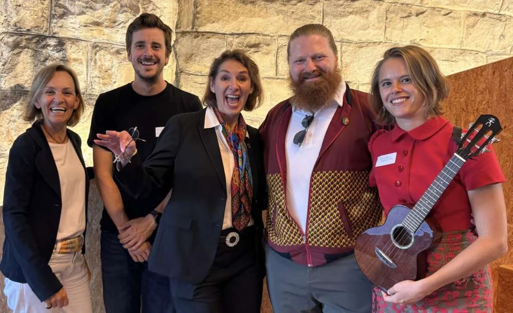 Five colleagues laughing together after a workshop, one holding a ukulele, against a sandstone wall