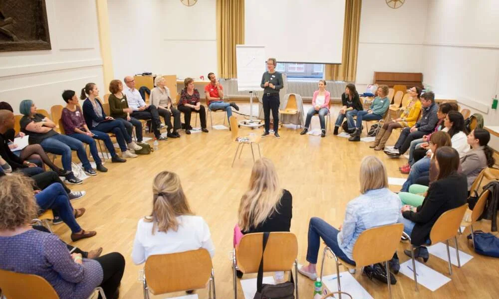 A facilitator speaking to a large circle of participants seated in a bright hall