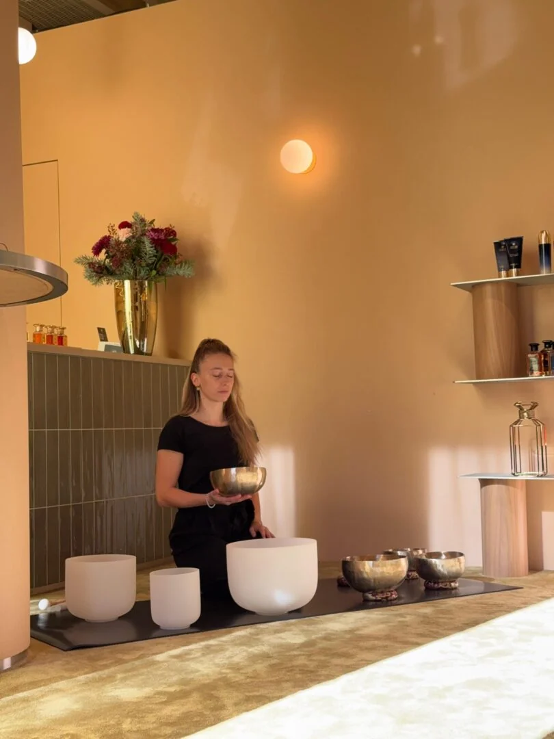 Céline playing a singing bowl beside a row of crystal bowls in a warm, softly lit room