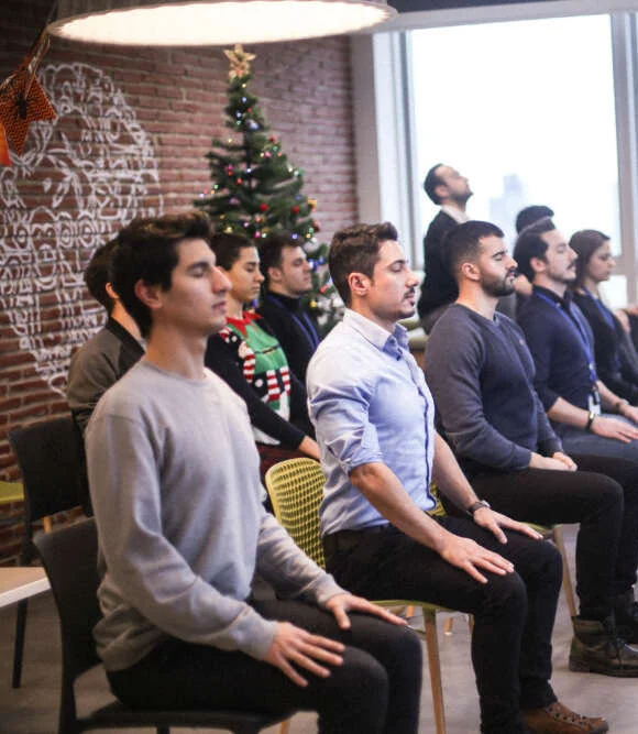 Colleagues sit upright on office chairs, eyes closed, during a guided mindfulness session by the window
