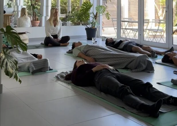 A teacher guiding a group resting on mats under blankets in a bright, plant-filled room