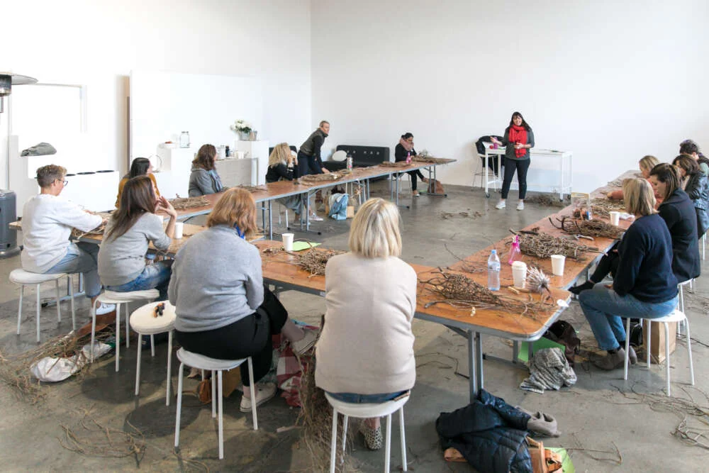 Colleagues at long tables working with bundles of foraged twigs while the artist speaks